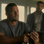 O Último Azul | Filme Brasileiro Leva o Urso de Prata no Festival de Berlim 2025 Alan Ritchson como Jack Reacher na terceira temporada de "The Reacher". Ele está sentado em uma mesa, vestido com camiseta lisa cinza e segurando um revólver com as duas mãos enquanto encara alguém do outro lado.