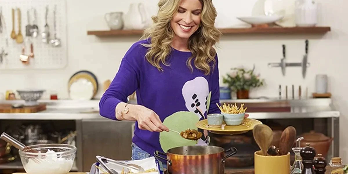 Rita Lobo em uma cozinha cheia, com uma blusa roxa, sorrindo, apresentando o "Cozinha Prática", do GNT.