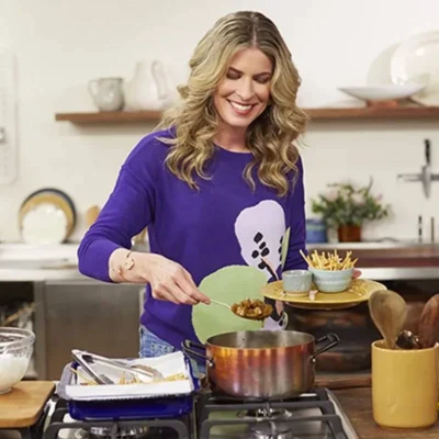 Rita Lobo Anuncia Saída do GNT Após 12 anos Para se Dedicar ao “Panelinha” Rita Lobo em uma cozinha cheia, com uma blusa roxa, sorrindo, apresentando o "Cozinha Prática", do GNT.