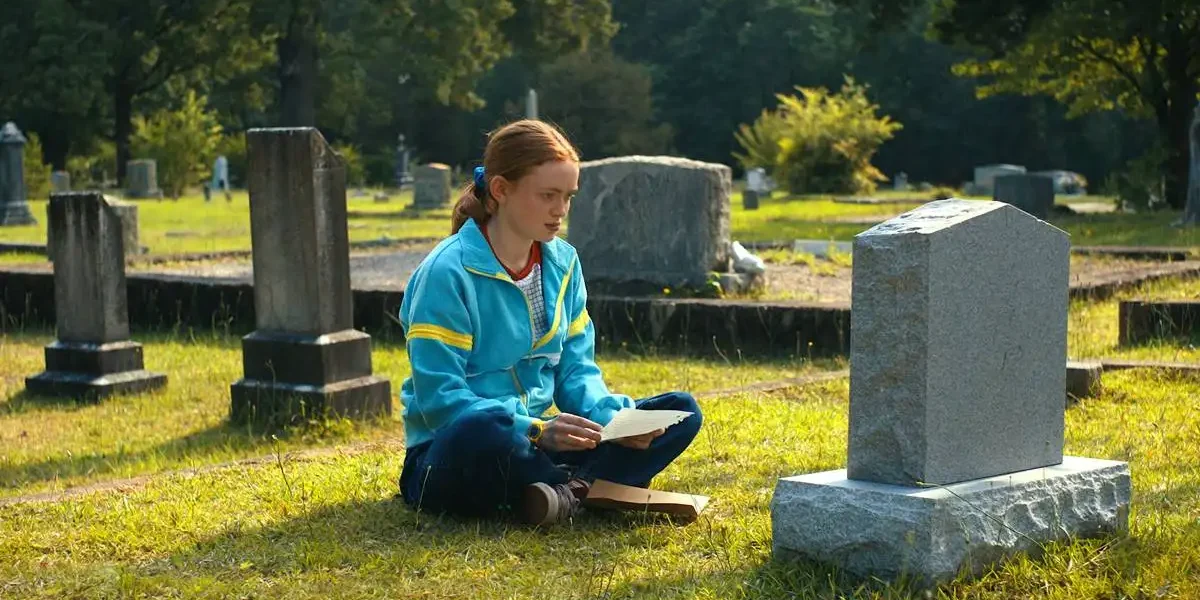 Sadie Sink como a adolescente Max, de "Stranger Things", quarta temporada. A garota ruiva está com um casaco azul e sentada de frente para o túmulo de seu irmão. Atriz está cotada para o novo X-Men.