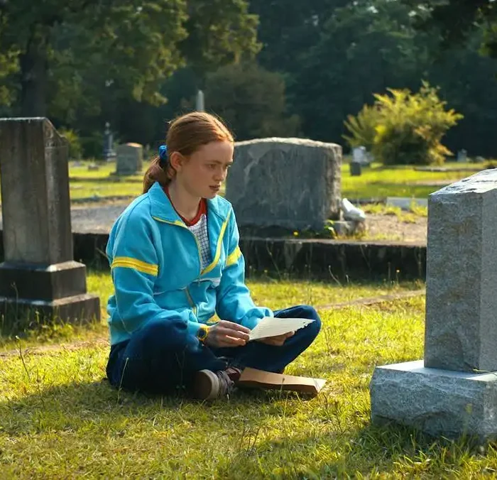 Sadie Sink como a adolescente Max, de "Stranger Things", quarta temporada. A garota ruiva está com um casaco azul e sentada de frente para o túmulo de seu irmão. Atriz está cotada para o novo X-Men.