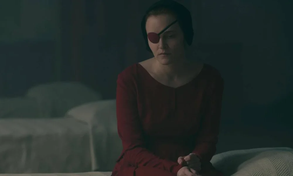 Imagem da aia Emily sentada em uma capa, com o traje vermelho e um tapa-olho. Imagem da 5ª temporada de "The Handmaid's Tale">