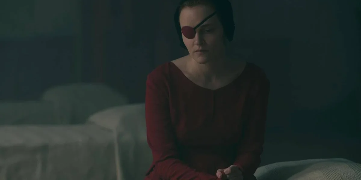 Imagem da aia Emily sentada em uma capa, com o traje vermelho e um tapa-olho. Imagem da 5ª temporada de "The Handmaid's Tale">