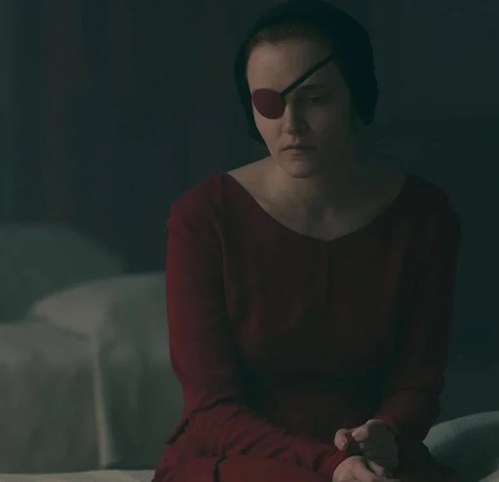 Imagem da aia Emily sentada em uma capa, com o traje vermelho e um tapa-olho. Imagem da 5ª temporada de "The Handmaid's Tale">