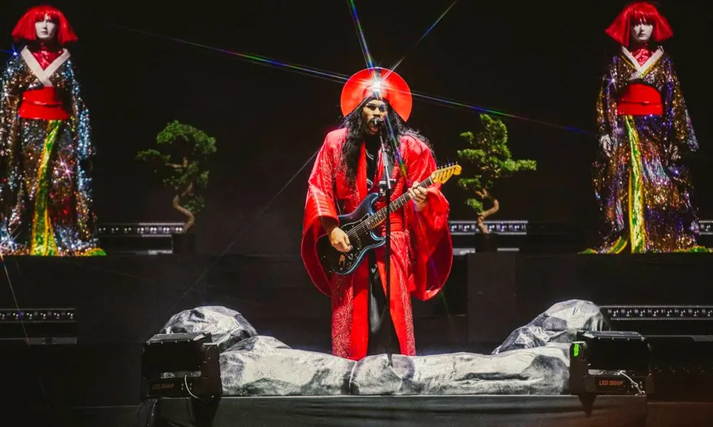 Luke Steele do Empire of The Sun, ao centro, com duas bailarinas ao seu lado ao fundo. Ele se veste com roupas místicas em conceito culto e intergalático, brilha uma luz refletindo de uma medalha no seu chapéu de semi-disco. As vestes são vermelhas e ele toca guitarra.