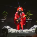 Citadel | Franquia da Série Tem Futuro Incerto no Prime Video Luke Steele do Empire of The Sun, ao centro, com duas bailarinas ao seu lado ao fundo. Ele se veste com roupas místicas em conceito culto e intergalático, brilha uma luz refletindo de uma medalha no seu chapéu de semi-disco. As vestes são vermelhas e ele toca guitarra.