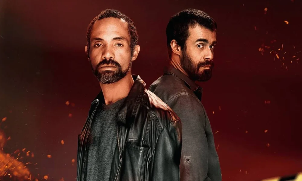 Atores Sílvio Guindane e Erom Cordeiro como os personagens Mendonça e Santiago da série "A Divisão", da Globoplay. Há um fundo vermelho com pontos de luz dando aparência de faíscas e incêndio surgindo da parte inferior da tela. Eles estão vestidos com jaquetas de couro e olham para a cãmera. Santiago, de costas para Mendonça, dá um olhar de canto.