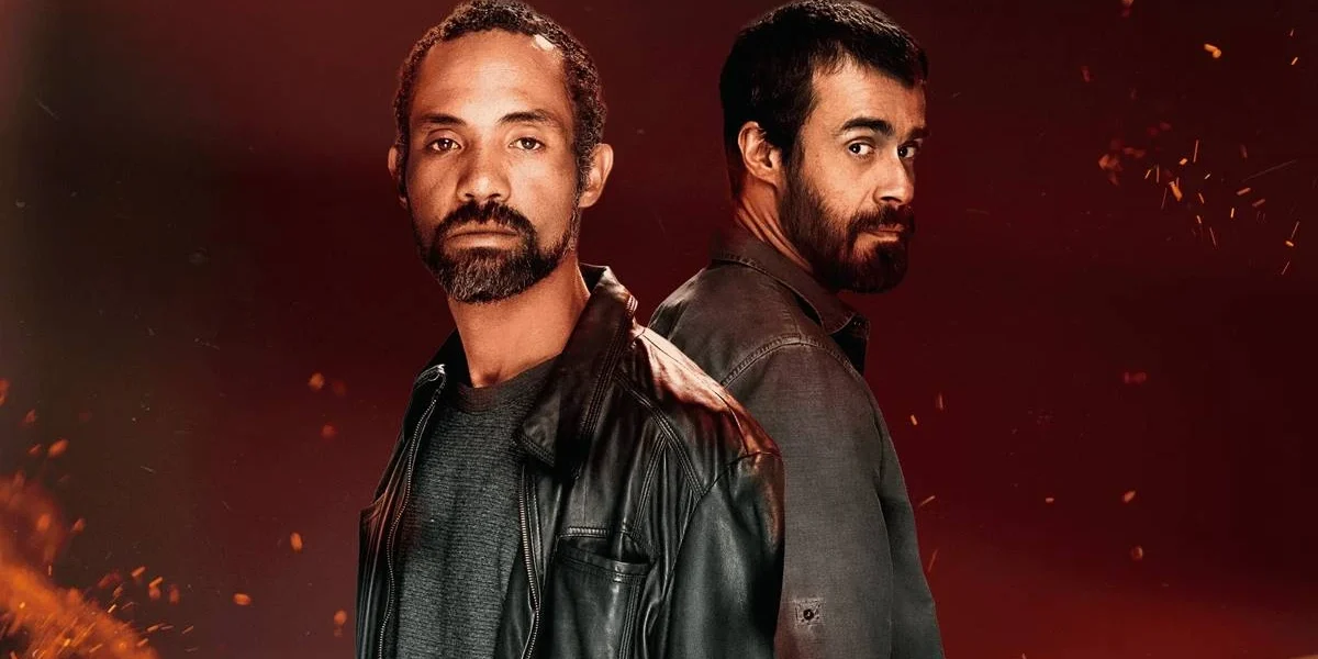 Atores Sílvio Guindane e Erom Cordeiro como os personagens Mendonça e Santiago da série "A Divisão", da Globoplay. Há um fundo vermelho com pontos de luz dando aparência de faíscas e incêndio surgindo da parte inferior da tela. Eles estão vestidos com jaquetas de couro e olham para a cãmera. Santiago, de costas para Mendonça, dá um olhar de canto.