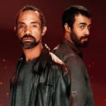 Atores Sílvio Guindane e Erom Cordeiro como os personagens Mendonça e Santiago da série "A Divisão", da Globoplay. Há um fundo vermelho com pontos de luz dando aparência de faíscas e incêndio surgindo da parte inferior da tela. Eles estão vestidos com jaquetas de couro e olham para a cãmera. Santiago, de costas para Mendonça, dá um olhar de canto.