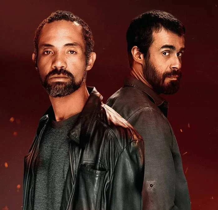 Atores Sílvio Guindane e Erom Cordeiro como os personagens Mendonça e Santiago da série "A Divisão", da Globoplay. Há um fundo vermelho com pontos de luz dando aparência de faíscas e incêndio surgindo da parte inferior da tela. Eles estão vestidos com jaquetas de couro e olham para a cãmera. Santiago, de costas para Mendonça, dá um olhar de canto.