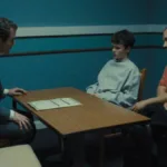 Sala de interrogatório para a série de drama "Adolescência", da Netflix. O rapaz de 13 anos está em destaque ao lado de seu pai, o interrogador está do outro lado da mesa, do lado esquerdo da tela.