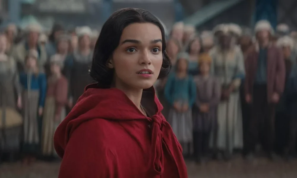 Rachel Zegler vestindo uma blusa com rapuz vermelho voltando ao reino do live-action de "Branca de Neve" (2025) interpretando a protagonista. A princesa está cercada por súditos ao fundo, desfocados da imagem.