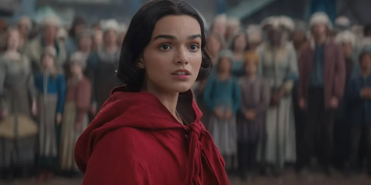Branca de Neve (2025) | Disney Divulga Trailer com Novas Imagens do Live-action Rachel Zegler vestindo uma blusa com rapuz vermelho voltando ao reino do live-action de "Branca de Neve" (2025) interpretando a protagonista. A princesa está cercada por súditos ao fundo, desfocados da imagem.