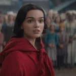 Rachel Zegler vestindo uma blusa com rapuz vermelho voltando ao reino do live-action de "Branca de Neve" (2025) interpretando a protagonista. A princesa está cercada por súditos ao fundo, desfocados da imagem.