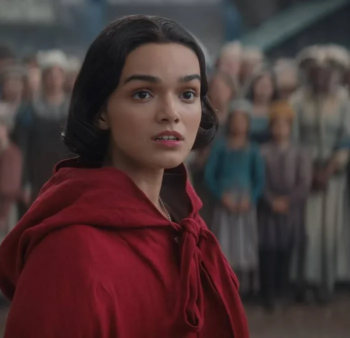 Rachel Zegler vestindo uma blusa com rapuz vermelho voltando ao reino do live-action de "Branca de Neve" (2025) interpretando a protagonista. A princesa está cercada por súditos ao fundo, desfocados da imagem.