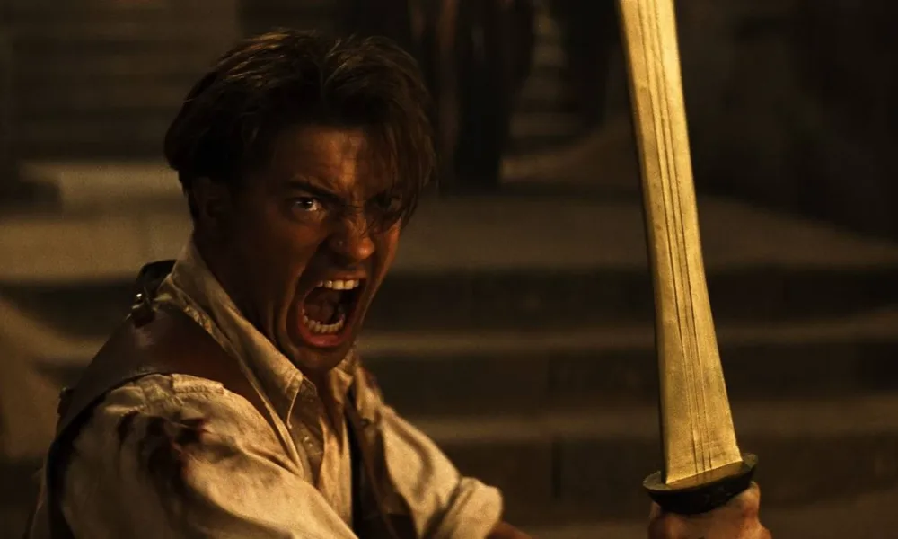 Brendan Fraser empunhando uma espada e gritando, dentro da tumba do filme "A Múmia", como o protagonista Rick O'Connell.