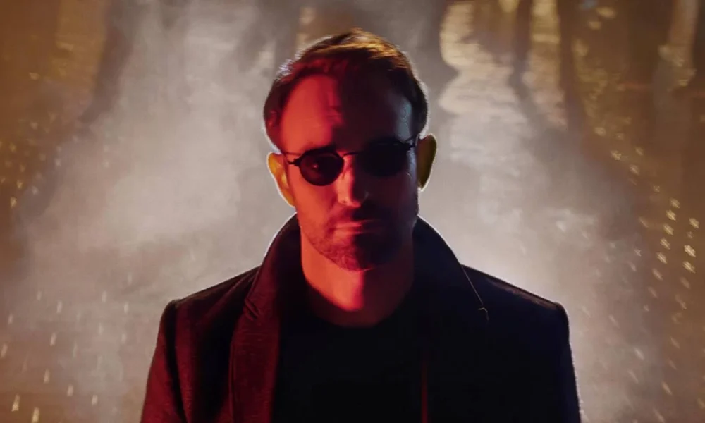 Protagonista de "Demolidor: Renascido" no primeiro episódio. Atrás dele uma explosão. Ele está de óculos e na sua cara há um filtro vermelho.