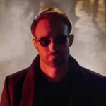 Protagonista de "Demolidor: Renascido" no primeiro episódio. Atrás dele uma explosão. Ele está de óculos e na sua cara há um filtro vermelho.