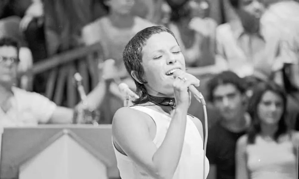 Elis Regina cantando, olhos fechados na imagem e um sorriso no rosto, no programa "Som Livre Exportação". Cantora está de cabelo curto, segurando o microfone de fio com a mão direita, e a imagem é em preto e branco.