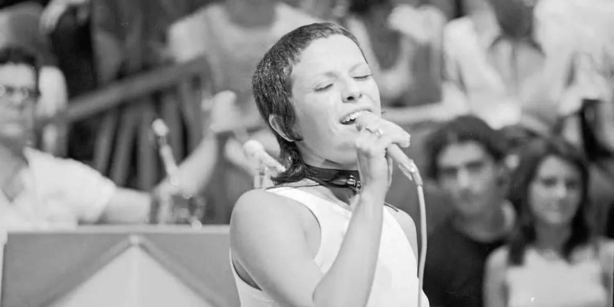 Elis Regina Faria 80 Anos Hoje e Segue Inesquecível Elis Regina cantando, olhos fechados na imagem e um sorriso no rosto, no programa "Som Livre Exportação". Cantora está de cabelo curto, segurando o microfone de fio com a mão direita, e a imagem é em preto e branco.