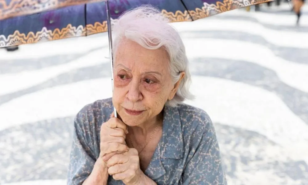 Fernanda Montenegro no calçadão do Rio de Janeiro com um guarda-sol, ela interpreta a protagonista do longa "Vitória" (2025) e olha distante. Seu cabelo parece estar sendo levado suavemente pelo vento à direita do telespectador; ela olha à esquerda.