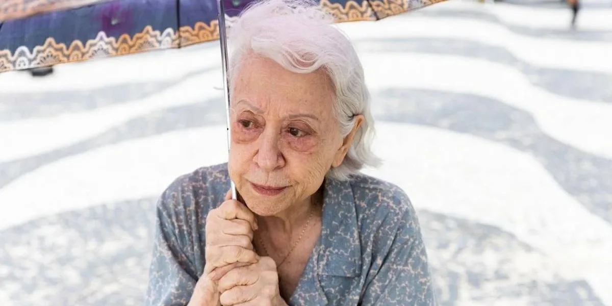 Fernanda Montenegro no calçadão do Rio de Janeiro com um guarda-sol, ela interpreta a protagonista do longa "Vitória" (2025) e olha distante. Seu cabelo parece estar sendo levado suavemente pelo vento à direita do telespectador; ela olha à esquerda.