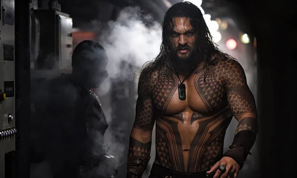 Jason Momoa como o Aquaman no filme homônimo de 2018. Ele está encarando um homem que está de costas para a câmera, há fumaça na tela, e podemos ver as tatuagens no tronco do personagem.