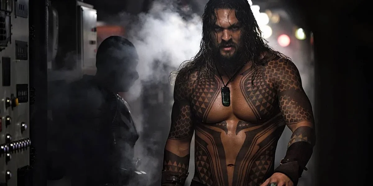 James Gunn Revela Pedido de Jason Momoa para Interpretar o Lobo Jason Momoa como o Aquaman no filme homônimo de 2018. Ele está encarando um homem que está de costas para a câmera, há fumaça na tela, e podemos ver as tatuagens no tronco do personagem.