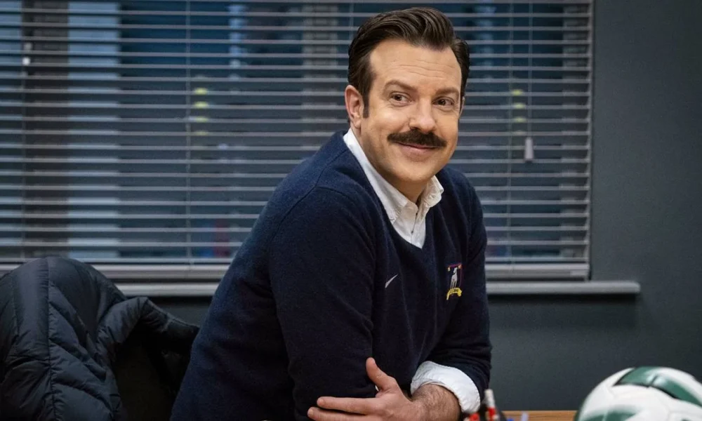 Jason sudeikis com um suéter interpretando o personagem Ted Lasso na série homônima, finale da temporada 3. Ele está apoiado em uma mesa e sorrindo.