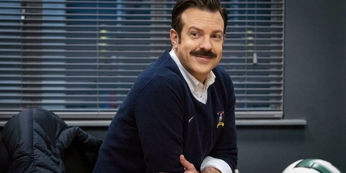 Jason sudeikis com um suéter interpretando o personagem Ted Lasso na série homônima, finale da temporada 3. Ele está apoiado em uma mesa e sorrindo.