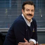 Jason sudeikis com um suéter interpretando o personagem Ted Lasso na série homônima, finale da temporada 3. Ele está apoiado em uma mesa e sorrindo.
