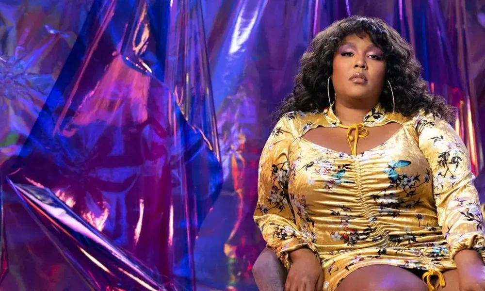 Lizzo para o reality "Watch Out For the Big Grrrls". Ela está na segunda metade de tela, com uma roupa amarela brilhante colada no corpo, maquiada com sombra púrpura, e o fundo parece um cristal roxo.