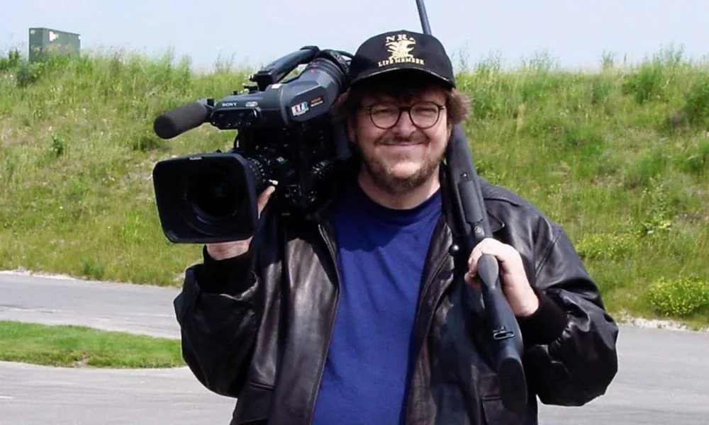 Michael Morre segurando uma câmera no ombro direito, vestindo uma camiseta azul e uma jaqueta preta por cima. Ele está de boné e sorrindo. Atrás dele há um campo verde com grama meio opaca. Imagem do documentário "Tiros em Columbine".