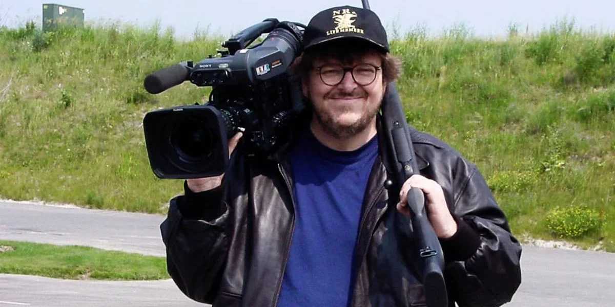 Michael Morre segurando uma câmera no ombro direito, vestindo uma camiseta azul e uma jaqueta preta por cima. Ele está de boné e sorrindo. Atrás dele há um campo verde com grama meio opaca. Imagem do documentário "Tiros em Columbine".