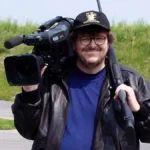 Michael Morre segurando uma câmera no ombro direito, vestindo uma camiseta azul e uma jaqueta preta por cima. Ele está de boné e sorrindo. Atrás dele há um campo verde com grama meio opaca. Imagem do documentário "Tiros em Columbine".