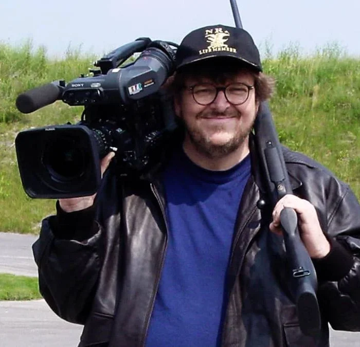 Michael Morre segurando uma câmera no ombro direito, vestindo uma camiseta azul e uma jaqueta preta por cima. Ele está de boné e sorrindo. Atrás dele há um campo verde com grama meio opaca. Imagem do documentário "Tiros em Columbine".
