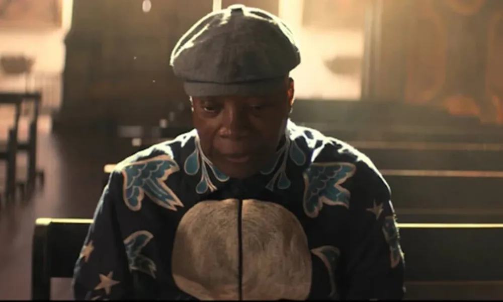 Milton Bituca Nascimento sentado, reflexivo, cabeça um pouco baixa, usando uma boina e roupa estampada escura com detalhes coloridos em azul e branco. O fundo é de uma igreja, na qual ele senta em um dos bancos.