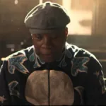 Milton Bituca Nascimento sentado, reflexivo, cabeça um pouco baixa, usando uma boina e roupa estampada escura com detalhes coloridos em azul e branco. O fundo é de uma igreja, na qual ele senta em um dos bancos.