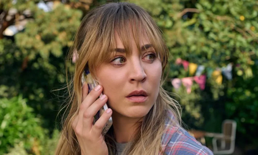 Kaley Cuoco como Emma, protagonista de "O Jogo do Disfarce" (2023), ou "Roleplay". Ela olha preocupada para a direita da tela, virando a cabeça, há árvores no fundo, e realiza uma ligação no telefone celular.