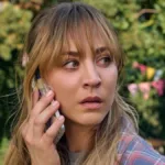 Kaley Cuoco como Emma, protagonista de "O Jogo do Disfarce" (2023), ou "Roleplay". Ela olha preocupada para a direita da tela, virando a cabeça, há árvores no fundo, e realiza uma ligação no telefone celular.