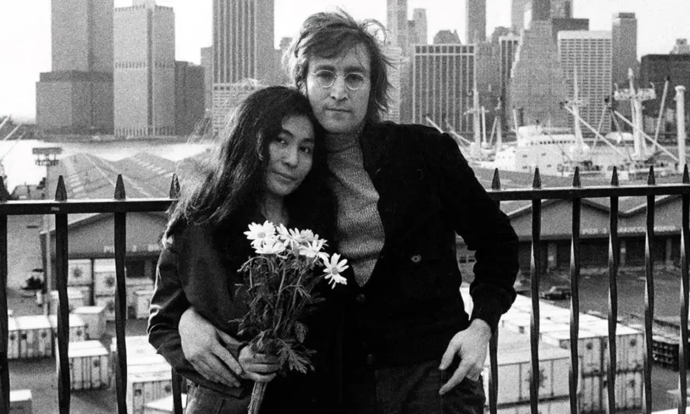 Yoko Ono, esquerda, e John Lennon, à direita, em fotografia em preto e branco. Eles recostam cabeças e ele a abraça por trás, com o corpo levemente inclinado. Ela segura um buquê do que parecem ser margaridas.