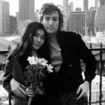 Yoko Ono, esquerda, e John Lennon, à direita, em fotografia em preto e branco. Eles recostam cabeças e ele a abraça por trás, com o corpo levemente inclinado. Ela segura um buquê do que parecem ser margaridas.