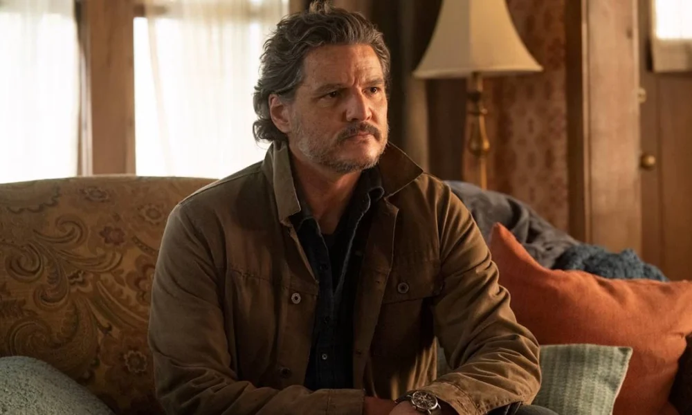 Pedro Pascal como Joel de "The Last of Us", na segunda temporada da série da HBO. Ele está em um sofá, olhando algo seriamente, e vestido com um casaco marrom.
