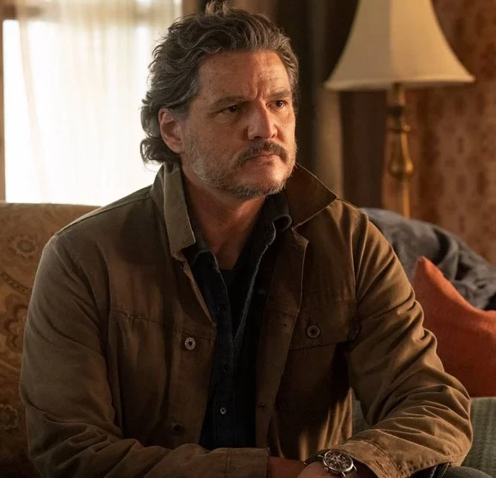 Pedro Pascal como Joel de "The Last of Us", na segunda temporada da série da HBO. Ele está em um sofá, olhando algo seriamente, e vestido com um casaco marrom.