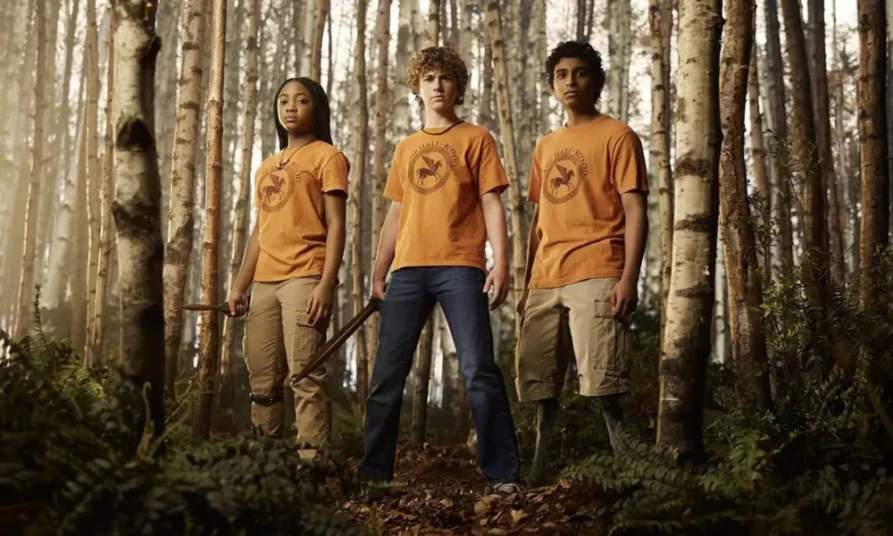 Annabeth, Percy, Grover, da série "Percy Jackson e os Olimpianos" em um bosque da série. Eles estão com o uniforme do acampamento e Percy está empunhando uma espada.