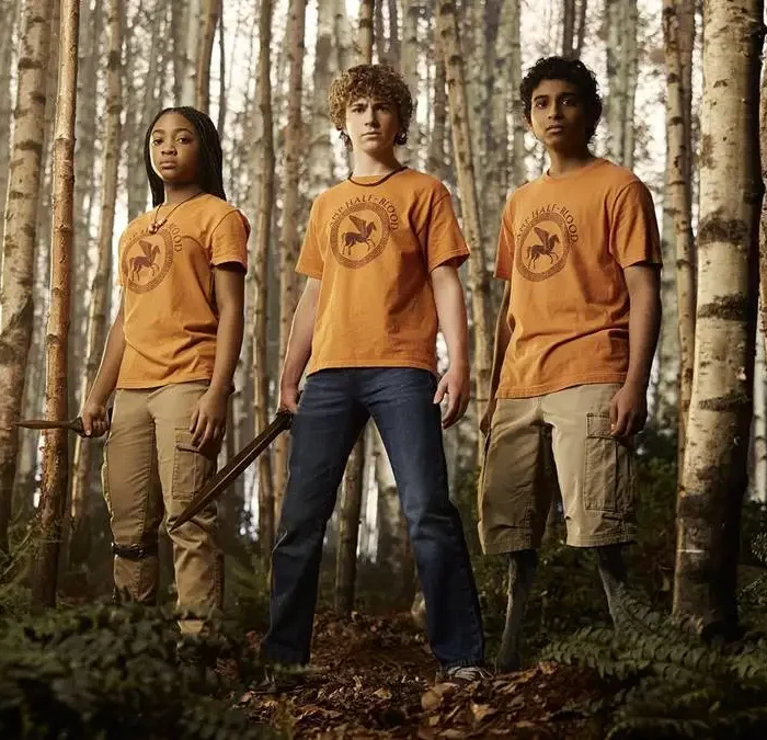 Annabeth, Percy, Grover, da série "Percy Jackson e os Olimpianos" em um bosque da série. Eles estão com o uniforme do acampamento e Percy está empunhando uma espada.