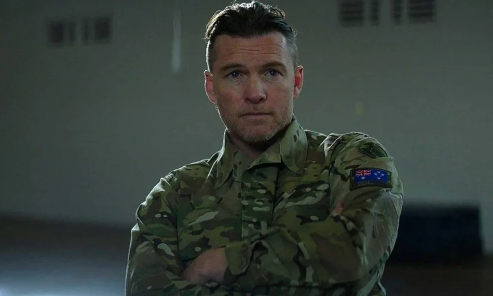 Sam Worthington como o protagonista de "Transfusão". Ele estácom uma cara neutra, olhando para um ponto perto da câmera, e vestido com trajes das forças armadas.