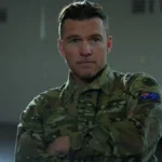 Paradise (2025) | Conspirações, Drama e Alteração da Realidade Sam Worthington como o protagonista de "Transfusão". Ele estácom uma cara neutra, olhando para um ponto perto da câmera, e vestido com trajes das forças armadas.