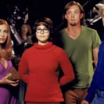 Daphone, Velma, Salsicha, Fred, nessa ordem, encarando alguém do "lado da câmera" no live-action de Scooby-Doo de 2002. Eles estão com as roupas tradicionais da série de animação e Daphne segura um prisma.