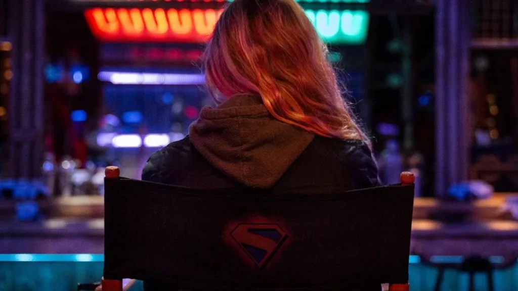 Supergirl | Nossas Primeiras Impressões 1 Atriz Milly Alcock sentada em uma cadeira de estúdio de gravações de cinema, de costas. Ela está em destaque e o fundo, a sua frente, está desfocado, e está com uma estética neon e cyberpunk vibrante. Supergirl